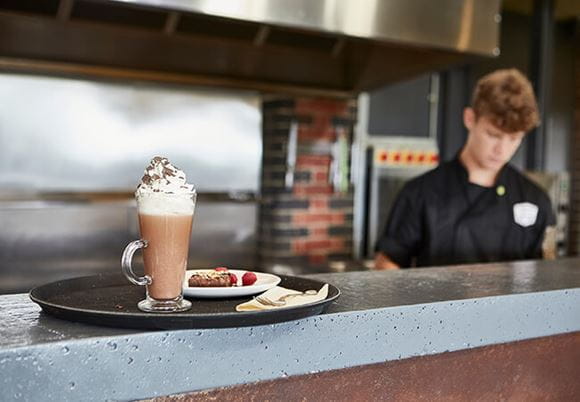 Kawa latte stojąca na blacie kawiarnianego baru. W tle pracujący kelner
